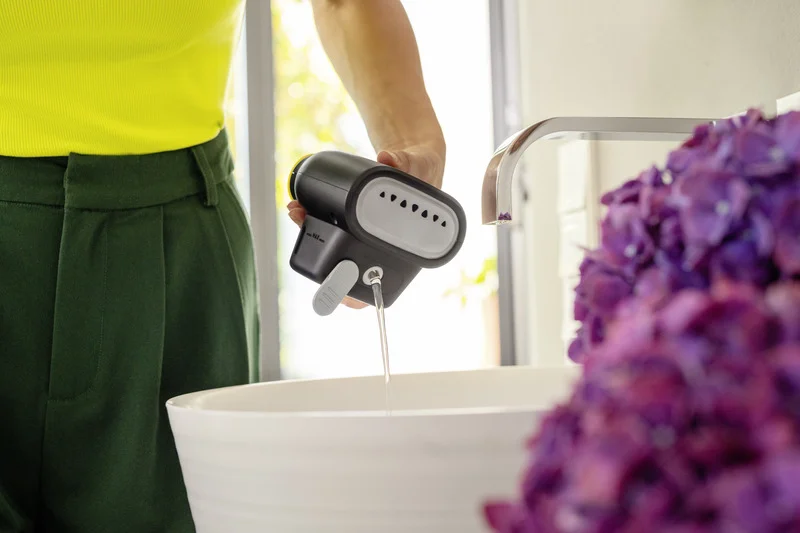 Person in gelbem Oberteil und grüner Hose hält einen Kärcher Dampfreiniger über einem weißen Waschbecken.
