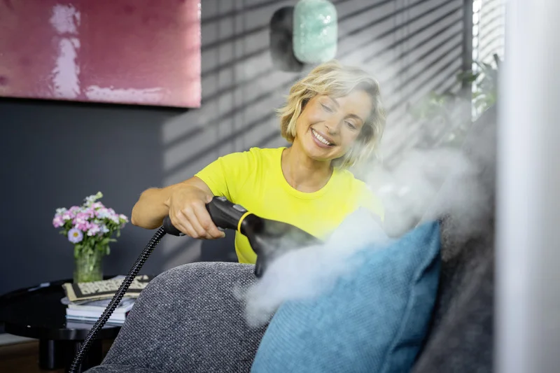 Person reinigt Sofa mit einem Kärcher Dampfreiniger in einem Wohnzimmer. Im Hintergrund sind Blumen und Bücher auf einem Tisch.