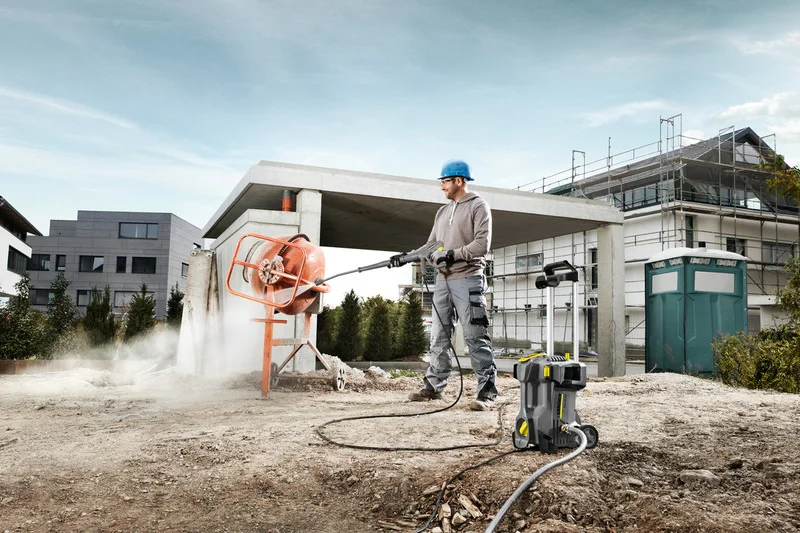 Bauarbeiter reinigt Betonmischer mit Kärcher Hochdruckreiniger auf Baustelle, umgeben von Gebäuden und Baugerüst.