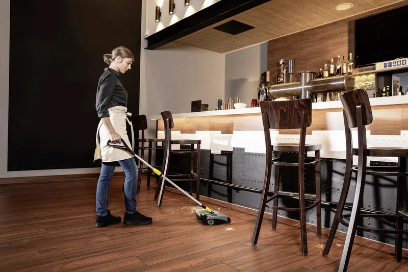 Person reinigt Holzboden in einer Bar mit einem Kärcher Akku-Besen. Hochstühle und Bar im Hintergrund.