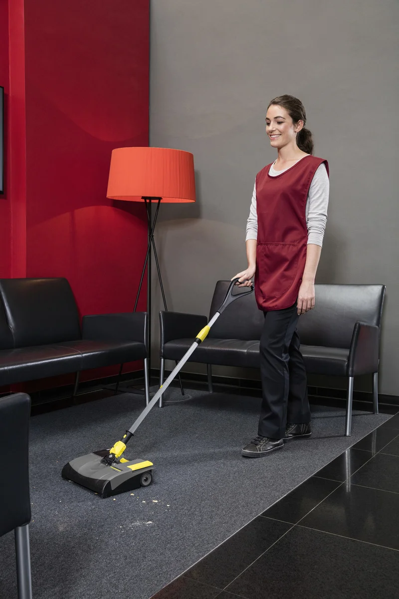 Person reinigt Teppichboden mit Kärcher Kehrmaschine in modernem Warteraum mit roten und grauen Wänden, schwarzen Ledersesseln und roter Stehlampe.