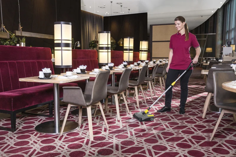 Frau reinigt Teppichboden in einem Restaurant mit einem Kärcher Kehrgerät. Umgebung mit roten Sitzbänken und gedeckten Tischen.