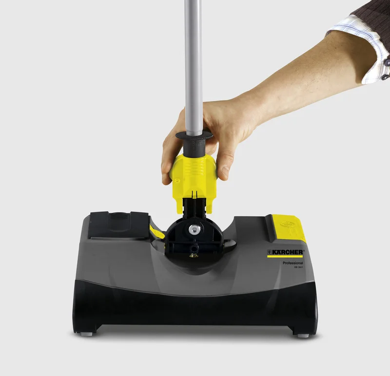Hand hält Griff eines Kärcher Kehrmaschinenaufsatzes. Produkt ist schwarz mit gelben Akzenten. Grauer Hintergrund.