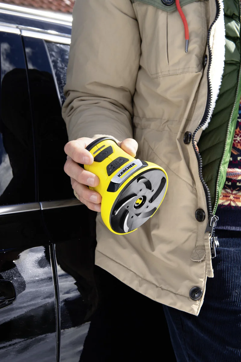 Person hält einen gelben Kärcher Polierer in der Hand, steht neben einem schwarzen Auto. Beige Jacke und bunter Pullover sichtbar.