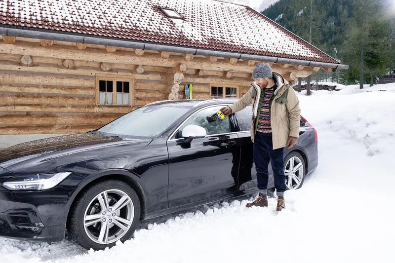 Person reinigt schwarze Limousine mit Kärcher Eiskratzer vor einer verschneiten Holzhütte im Freien. Skier lehnen an der Hütte.