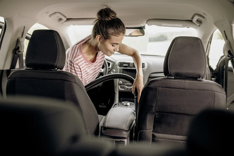 Person reinigt den Innenraum eines Autos mit einem Staubsauger. Sie trägt ein gestreiftes T-Shirt und beugt sich über die Mittelkonsole.