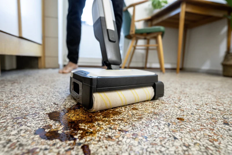 Kärcher Bodenreiniger entfernt Flüssigkeit von einem Steinboden in einem Wohnzimmer. Person im Hintergrund.
