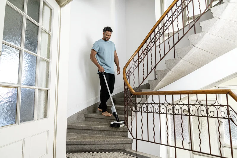 Person reinigt Treppe mit Kärcher Akku-Besen in einem hellen Treppenhaus mit Metallgeländer und Glastür.