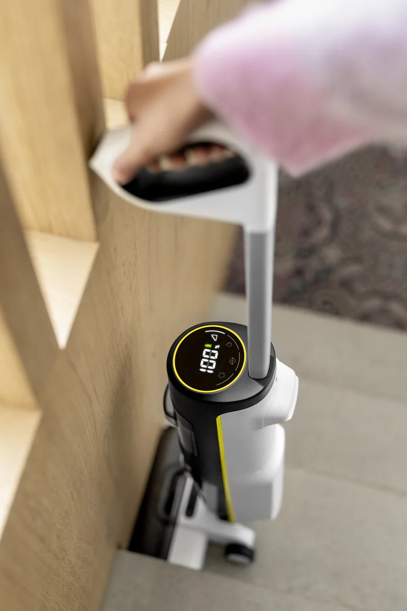 Hand hält Kärcher Akku-Bodenreiniger an Treppe. Gerät zeigt digitale Anzeige mit Zahlen und Symbolen. Holzgeländer im Hintergrund.