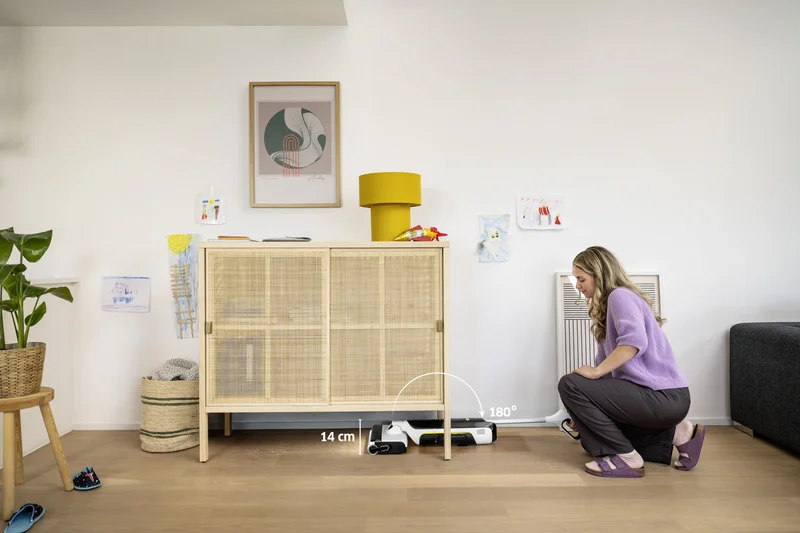 Frau hockt neben einem Kärcher Saugroboter, der unter einem Holzschrank in einem modernen Wohnzimmer steht. Eine Grafik zeigt eine Messung von 34 cm.