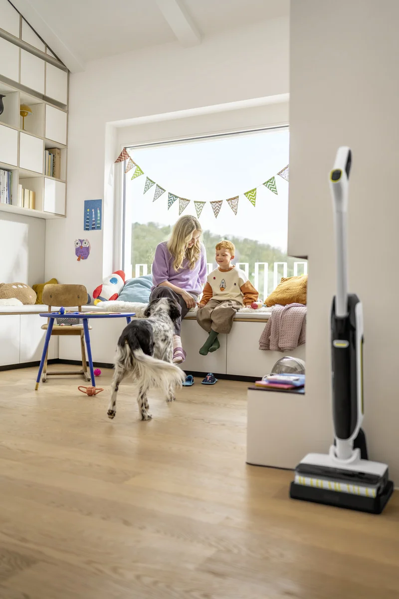 Frau, Kind und Hund in einem hellen Raum mit Holzboden. Sie sitzen auf einer Fensterbank mit Wimpelkette. Im Vordergrund steht ein Kärcher Hartbodenreiniger.