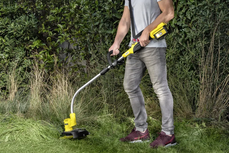 Mann bedient Kärcher Rasentrimmer im Garten, trägt graues T-Shirt und Jeans, umgeben von Gras und Sträuchern.