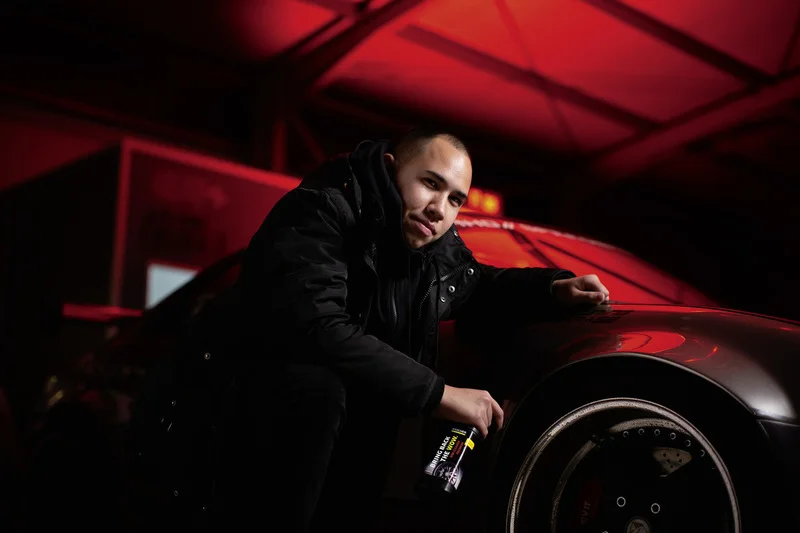Person in schwarzer Jacke kniet neben einem Auto mit roten Lichtern, hält eine Flasche "Race Glaze" in der Hand.