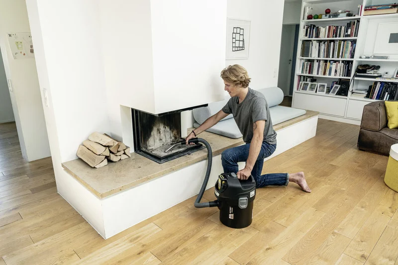 Person reinigt Kamin mit Kärcher Aschesauger in modernem Wohnzimmer, Holzscheite neben Kamin, Bücherregal im Hintergrund.