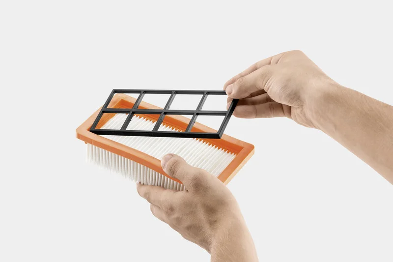 Zwei Hände halten einen rechteckigen Luftfilter mit orangefarbenem Rahmen und schwarzem Gitter auf weißem Hintergrund.