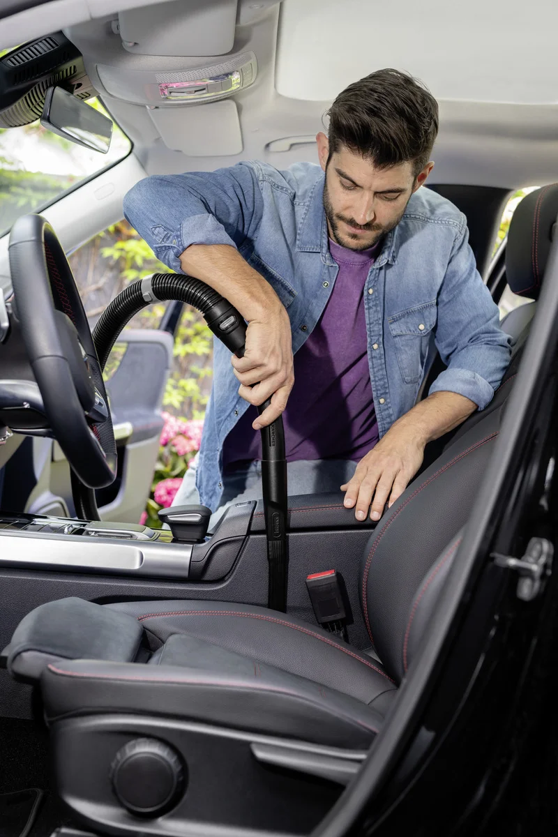 Mann reinigt Autoinnenraum mit einem Kärcher Staubsauger. Er trägt ein Jeanshemd und steht neben dem geöffneten Autotür.