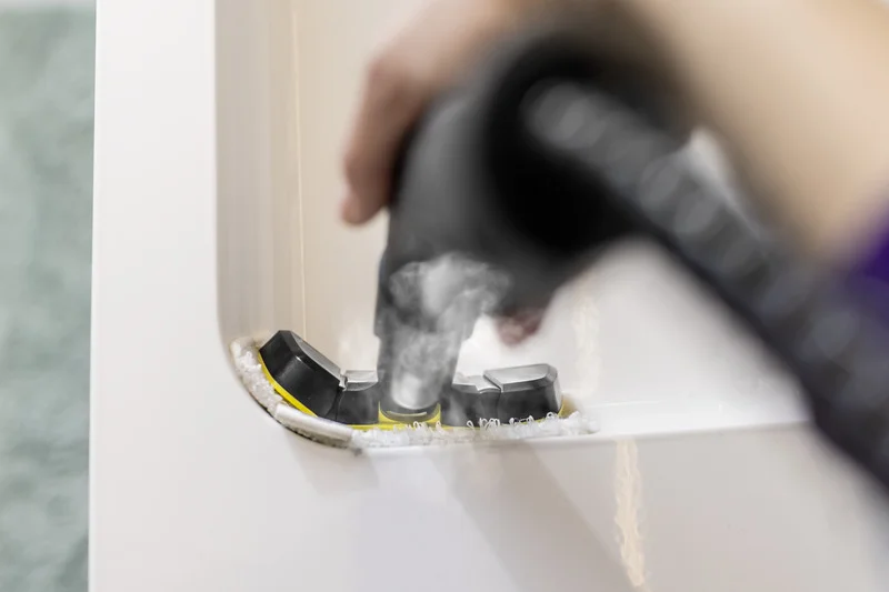 Hand hält Kärcher Dampfreiniger, reinigt weiße Fliesenfuge mit Bürstenaufsatz. Dampf sichtbar. Indoor-Szene.