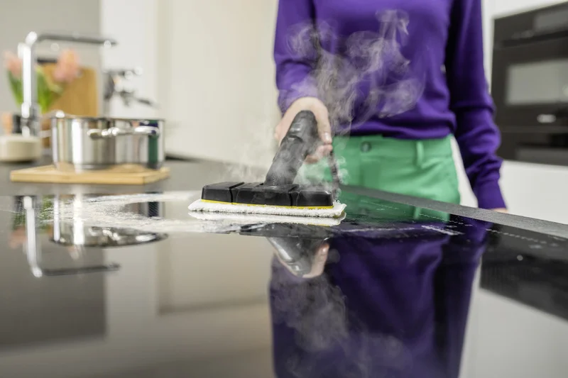 Person reinigt mit Kärcher Dampfreiniger eine schwarze Küchenarbeitsplatte, Dampf sichtbar, Topf im Hintergrund.