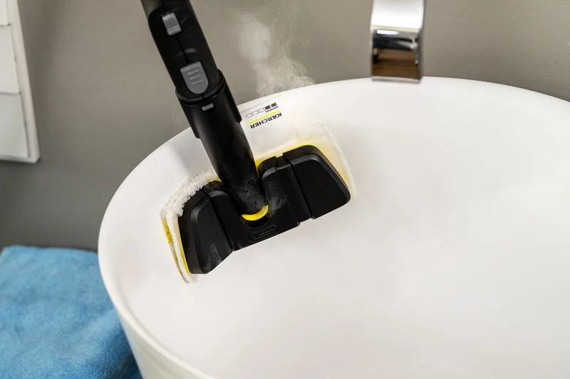 Kärcher Dampfreiniger reinigt ein weißes Waschbecken, Dampf steigt auf, daneben liegt ein blaues Handtuch.
