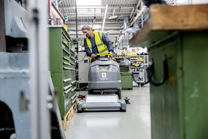 Person in gelber Warnweste bedient Kärcher Bodenreinigungsmaschine in einer Werkstatt mit grünen Werkzeugschränken und industrieller Ausstattung.