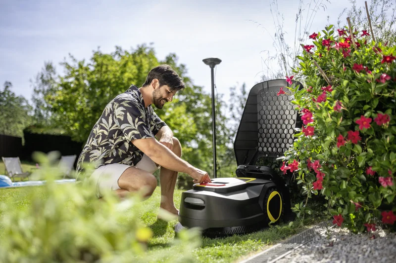 Mann kniet im Garten neben einem Kärcher Mähroboter, umgeben von blühenden roten Blumen und grünem Rasen.