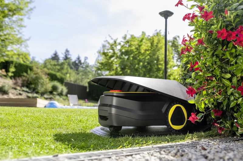 Kärcher Mähroboter in einem Garten neben blühenden roten Blumen, im Hintergrund Bäume und ein Zaun.