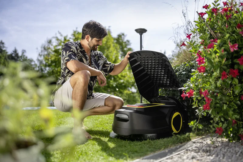 Person kniet im Garten neben einem geöffneten Kärcher Mähroboter, umgeben von blühenden Pflanzen und Bäumen.