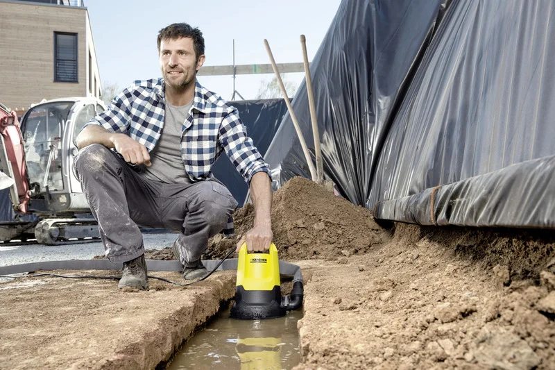 Mann kniet neben einem Graben mit Wasser und hält eine gelbe Kärcher Tauchpumpe. Baustelle im Hintergrund mit Bagger und Gebäude.