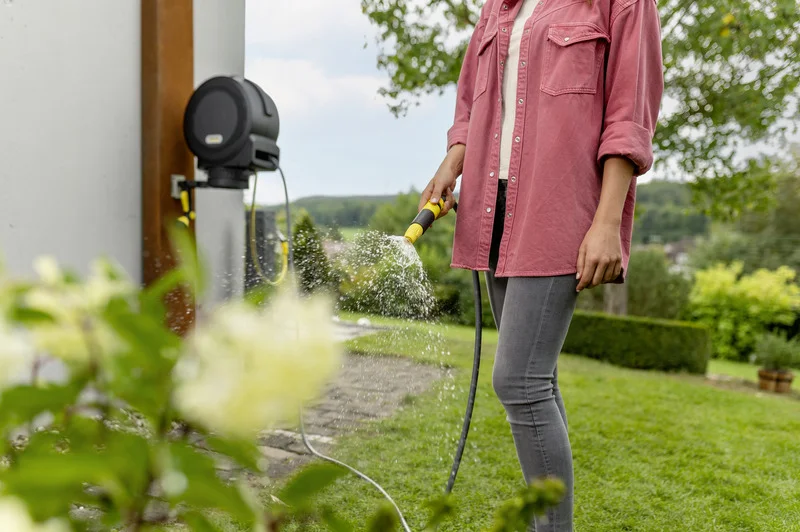 Person in rosa Hemd bewässert Garten mit Kärcher Gartenschlauch. Im Hintergrund grüne Landschaft und Pflanzen.