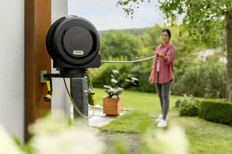Kärcher Schlauchbox an einer Wand im Garten, Person hält den Schlauch. Im Hintergrund grüne Pflanzen und Bäume.