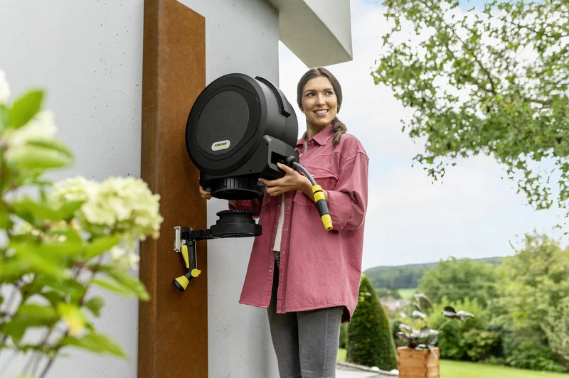 Person montiert Kärcher Schlauchaufroller an einer Wand im Freien, umgeben von Pflanzen und einem Garten im Hintergrund.