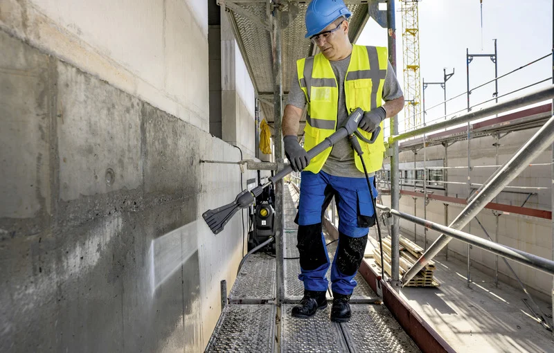 Arbeiter in Sicherheitskleidung reinigt Betonwand mit Kärcher Hochdruckreiniger auf Baustelle. Gerüst und Kran im Hintergrund.