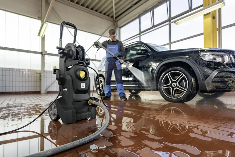 Person reinigt schwarzes Auto mit Kärcher Hochdruckreiniger in heller Werkstatt, Wasser spritzt auf nassen Boden.