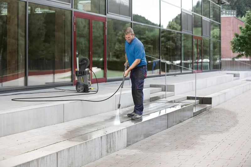 Mann reinigt Außentreppe mit Kärcher Hochdruckreiniger vor einem modernen Gebäude mit großen Glasfenstern.