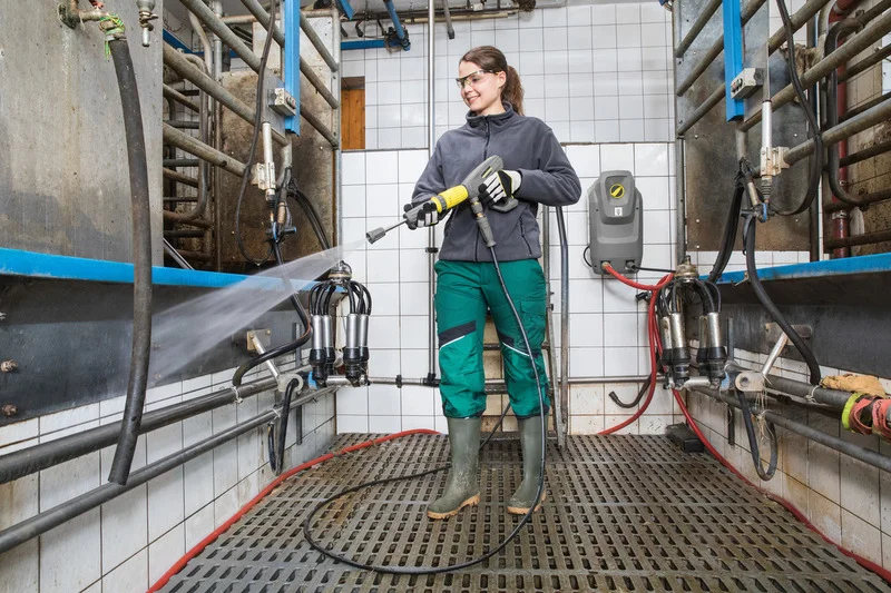 Person reinigt mit Kärcher Hochdruckreiniger eine Melkmaschine in einem gefliesten Raum. Umgebung mit Metallrohren und Gitterboden.