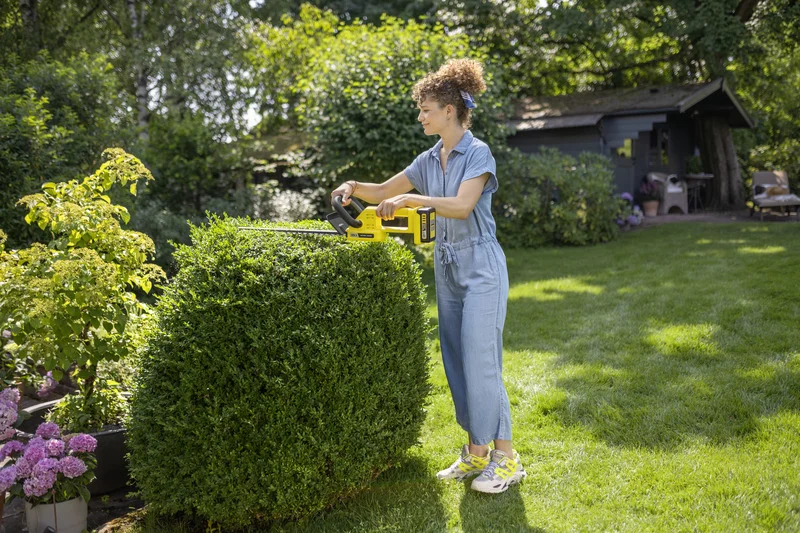 Frau schneidet Hecke im Garten mit einem Kärcher Heckenschneider. Im Hintergrund sind Bäume und ein Gartenhaus sichtbar.
