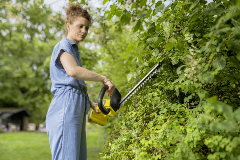 Person in blauem Overall schneidet mit einem Kärcher Heckenschere eine grüne Hecke im Freien.