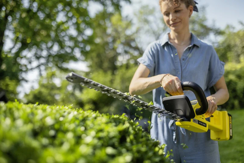 Person schneidet Hecke mit einer gelben Kärcher Heckenschere im Garten. Sonniges Wetter, grüne Pflanzen im Hintergrund.