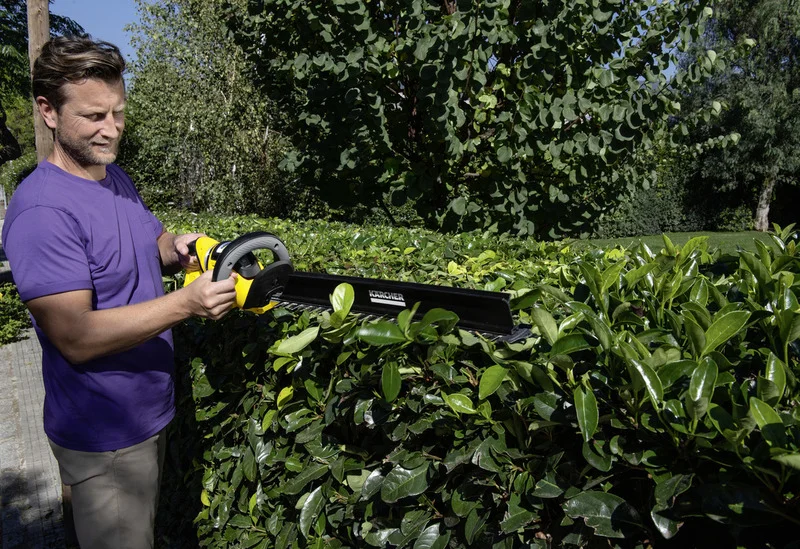 Person schneidet Hecke mit Kärcher Heckenschere im Garten. Umgeben von grünen Bäumen und sonnigem Wetter.