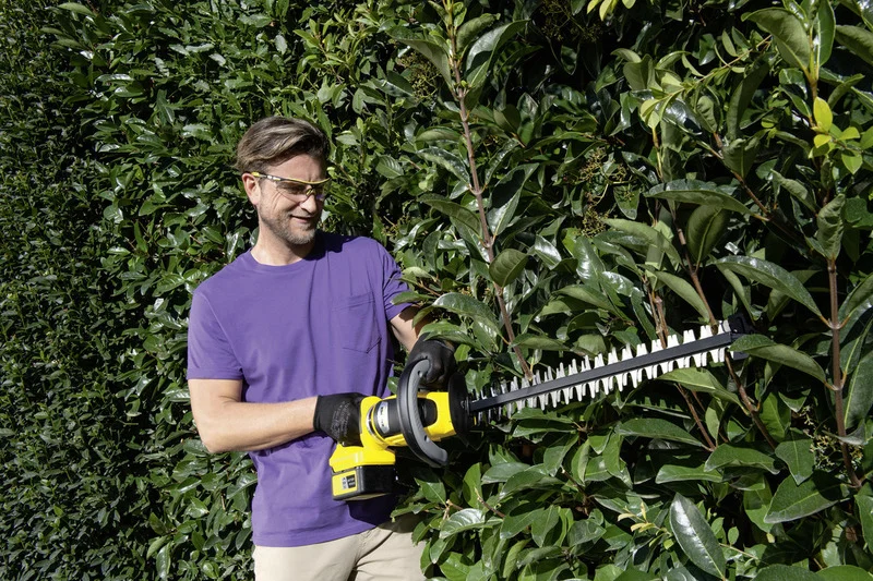 Person in lila T-Shirt benutzt eine gelbe Kärcher Heckenschere, um eine grüne Hecke im Freien zu trimmen.