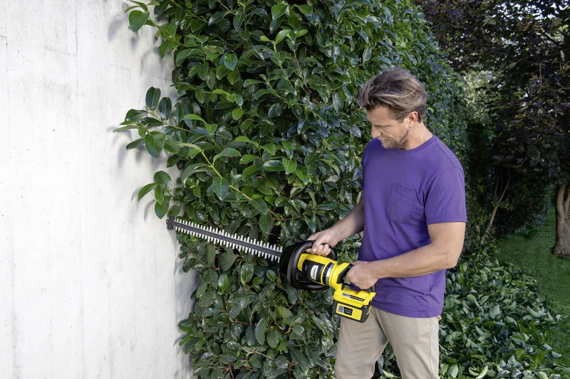 Person schneidet Hecke mit Kärcher Heckenschere im Garten. Die Hecke wächst an einer weißen Wand entlang.