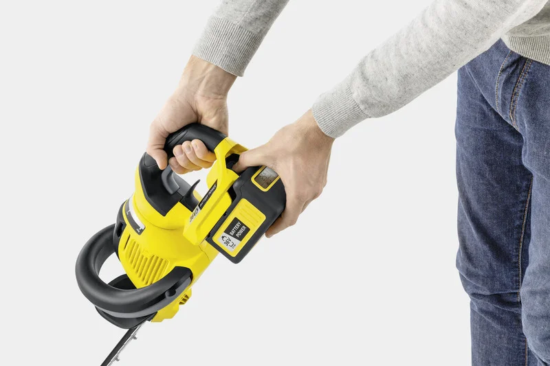 Person hält gelben Kärcher Akku-Heckenschere mit beiden Händen, grauer Pullover und blaue Jeans sichtbar.