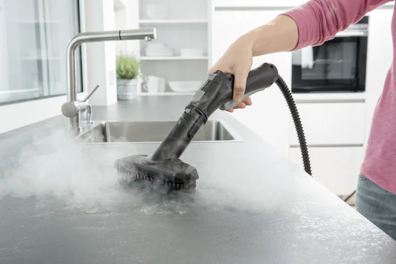 Hand hält Kärcher Dampfreiniger über Küchentheke, Dampf steigt auf, Spüle und Geschirr im Hintergrund.