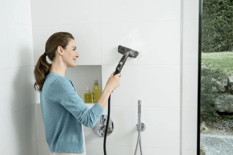 Person reinigt Badezimmerfliesen mit einem Kärcher Dampfreiniger. Im Hintergrund sind Shampoo-Flaschen und eine Dusche sichtbar.