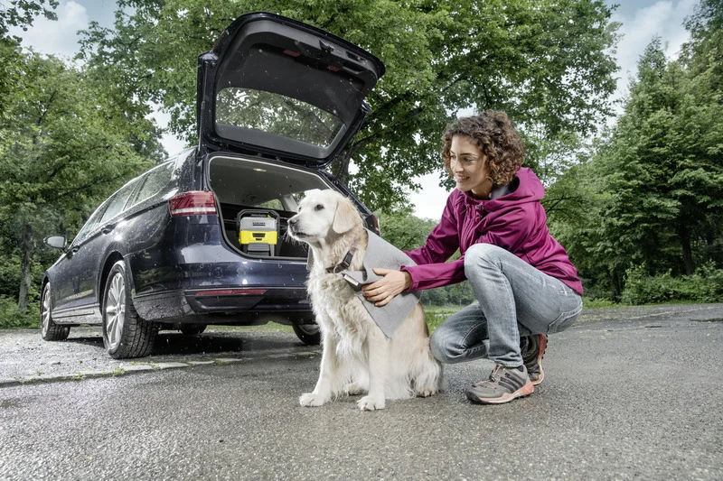 Frau kniet neben einem Hund auf einem Parkplatz. Im geöffneten Kofferraum eines Autos steht ein Kärcher Hochdruckreiniger.