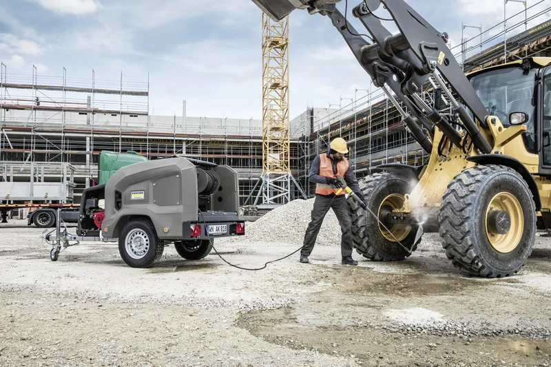 Bauarbeiter reinigt Rad eines Baggers mit Kärcher Hochdruckreiniger auf Baustelle. Im Hintergrund Baugerüst und Kran.
