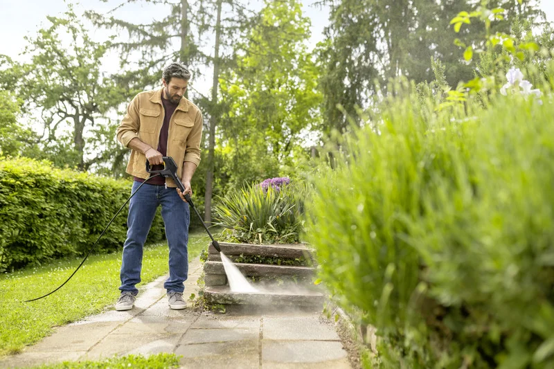 Person reinigt Außentreppe mit Kärcher Hochdruckreiniger im Garten. Umgeben von grünen Pflanzen und Bäumen.