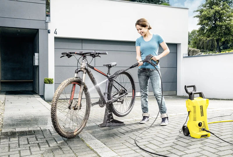 Frau reinigt ein Mountainbike mit einem Kärcher Hochdruckreiniger auf einer gepflasterten Auffahrt vor einer modernen Garage.