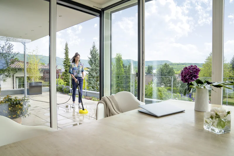 Person reinigt Terrasse mit Kärcher Hochdruckreiniger. Innenraum mit Tisch, Laptop und Blumen. Blick auf Garten und Berge durch Fenster.