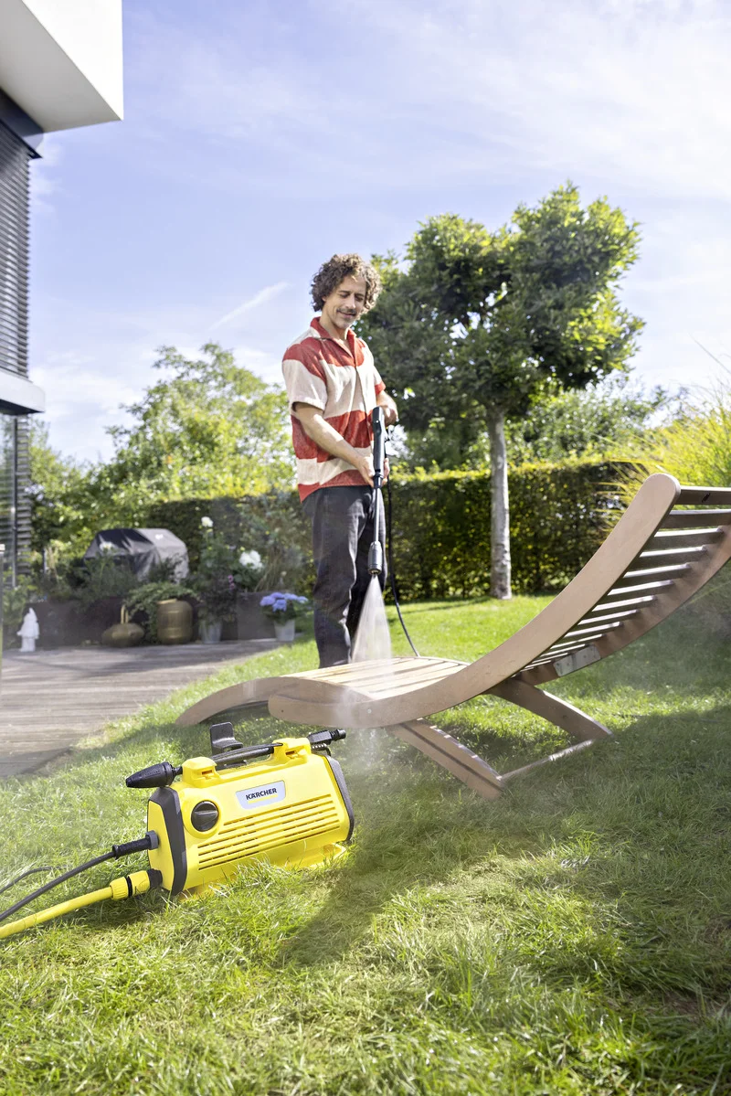 Person reinigt Gartenliege mit Kärcher Hochdruckreiniger im Freien. Im Hintergrund sind Bäume und ein Haus sichtbar.